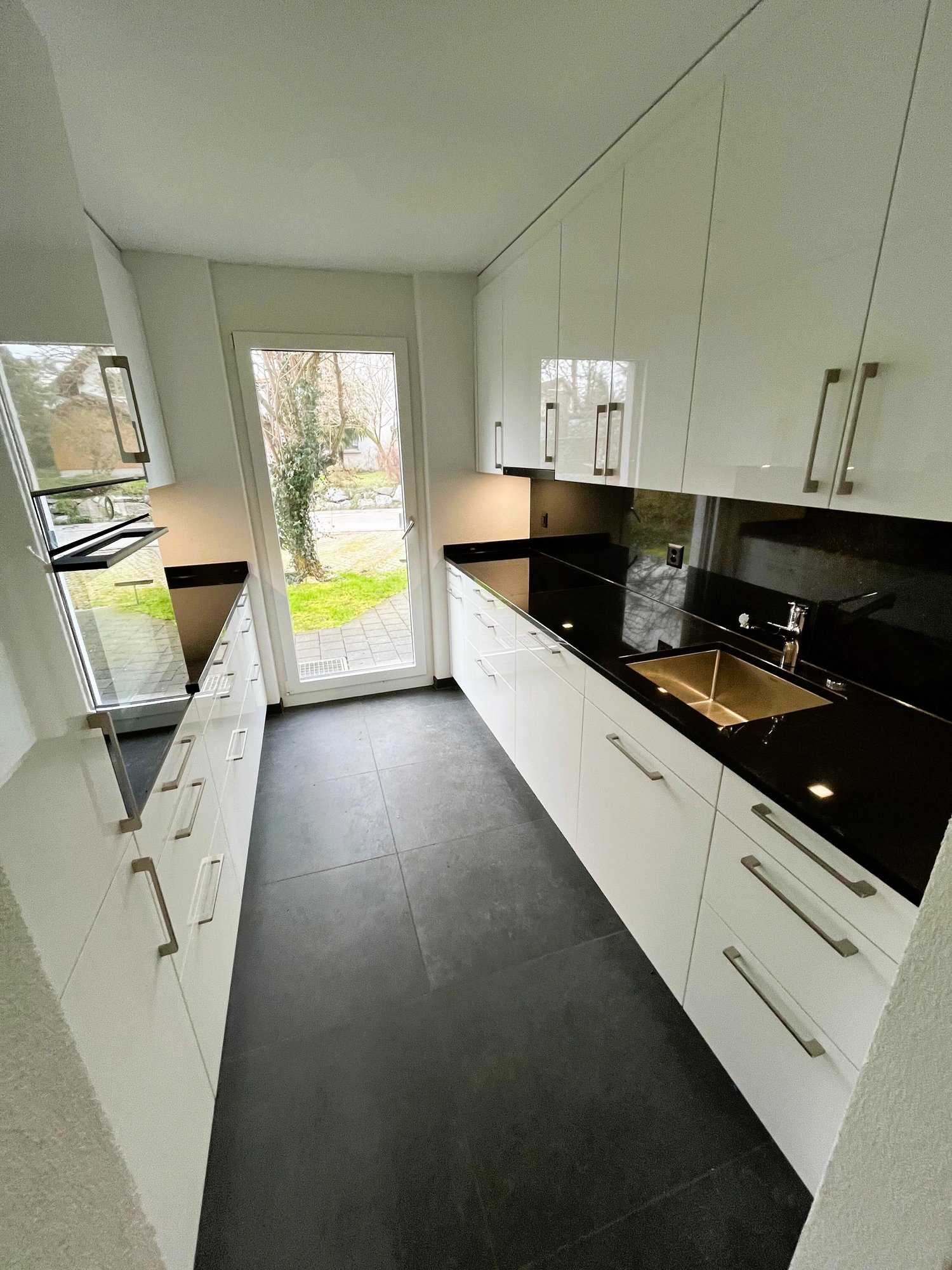 Modern, minimalist kitchen with white cabinets, black countertops, stainless steel appliances, and a large window overlooking the outdoor garden area.