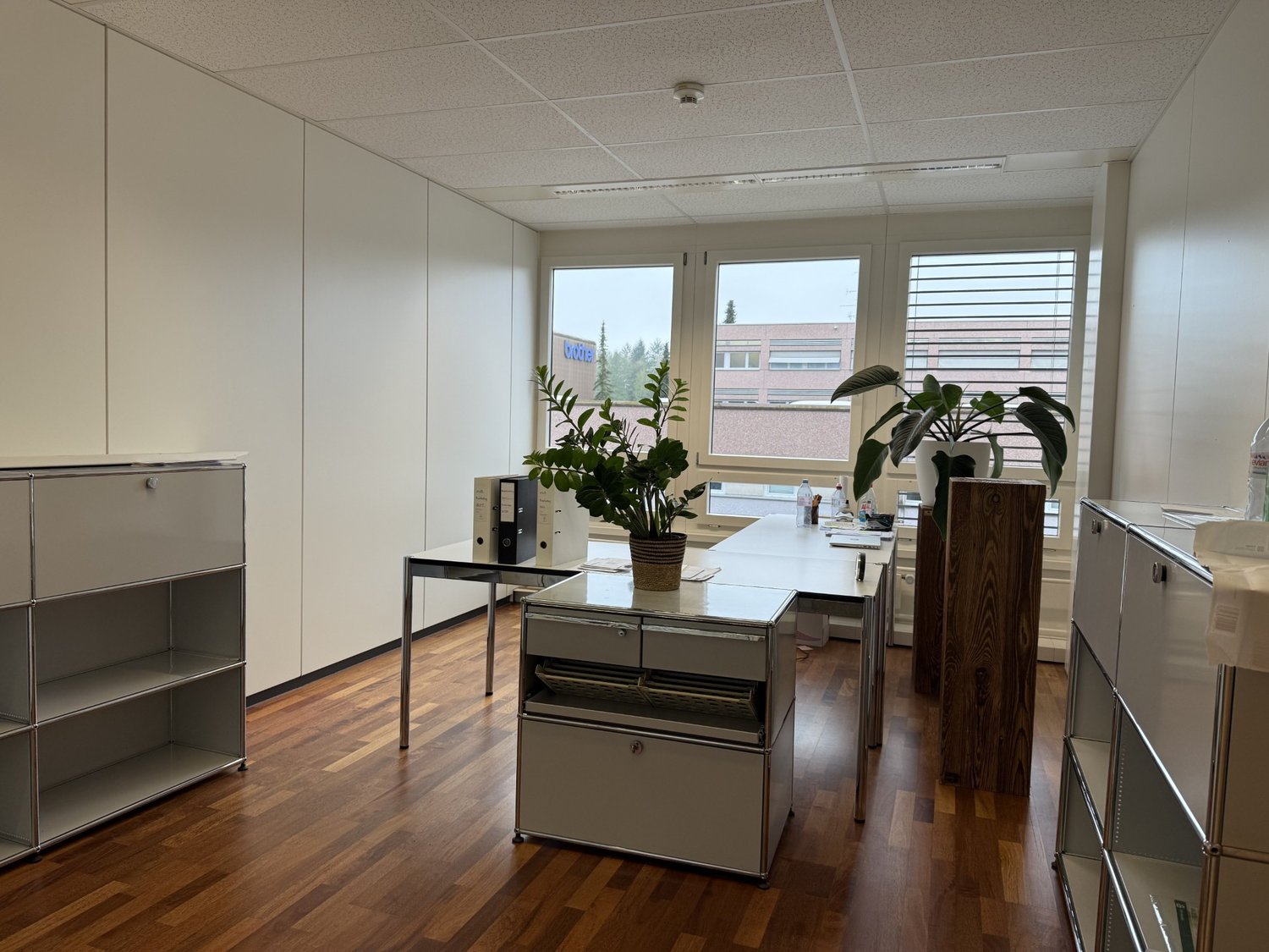An empty office with white walls, wooden floor, white cabinets, two desks, and two potted plants.