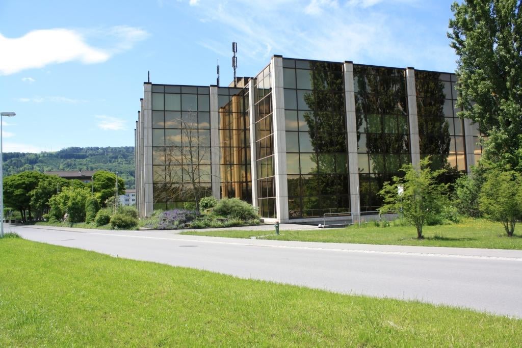 Modern office building, glass facade, multiple floors, surrounded by greenery, road in front