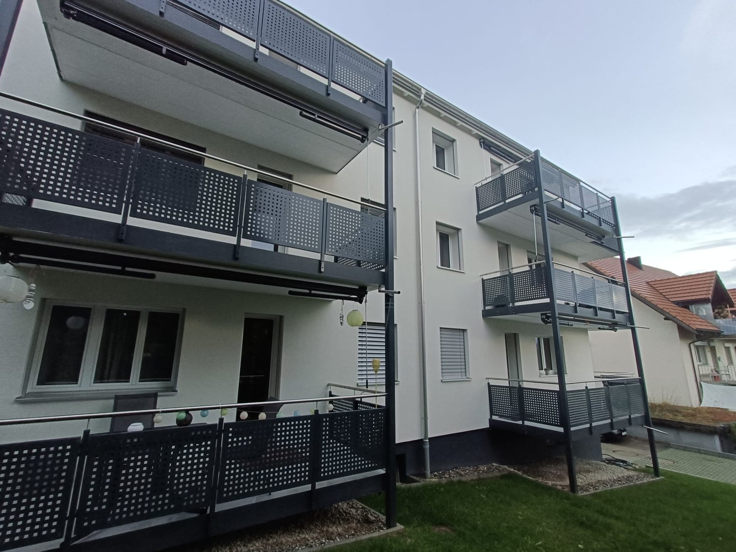 Modern apartment building with multiple balconies, white exterior, black railings, and a small garden in front.