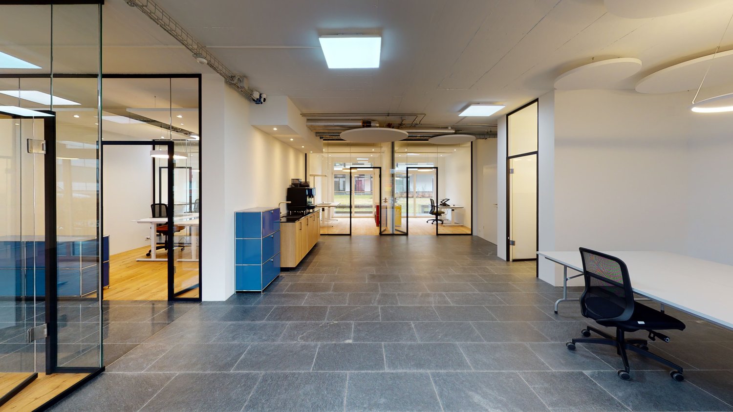 Open space office with glass walls, wooden floors, white walls, modern furniture, and ceiling lights.