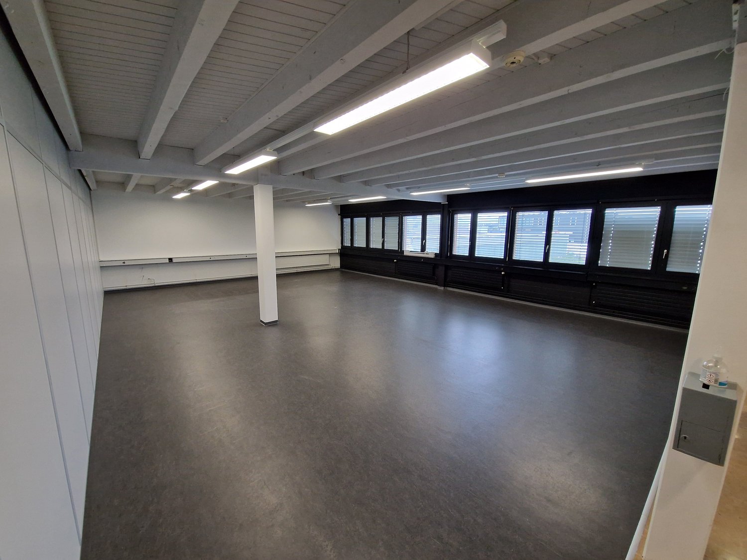 Empty room with white walls, gray floor, white ceiling with beams, large windows, and fluorescent lights