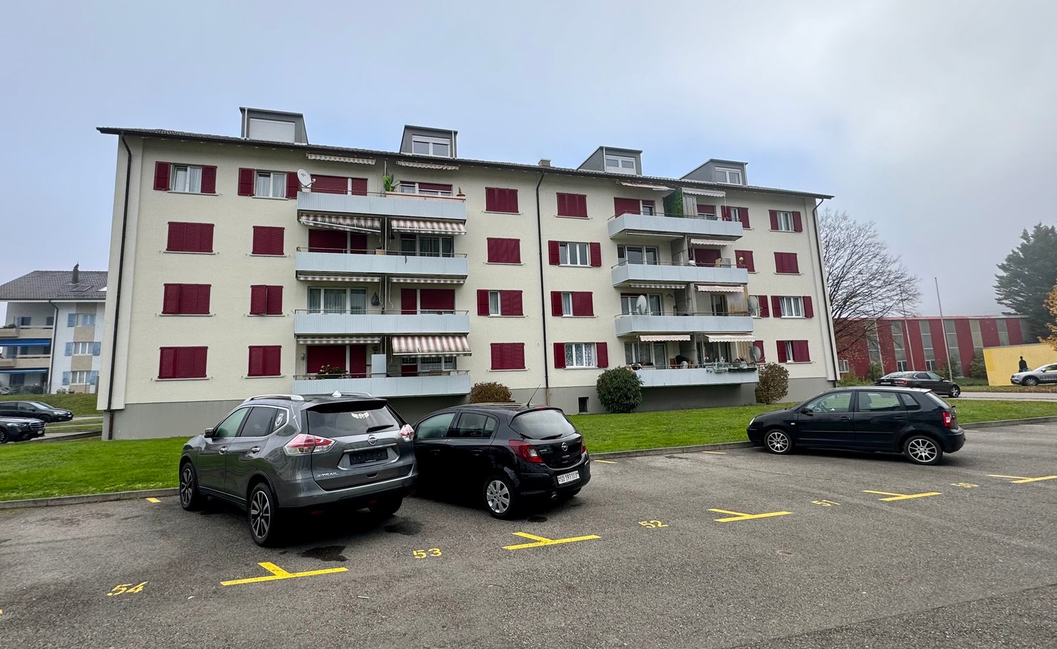 White and red apartment building with multiple balconies, cars parked in front, grassy area with a yellow building in the background
