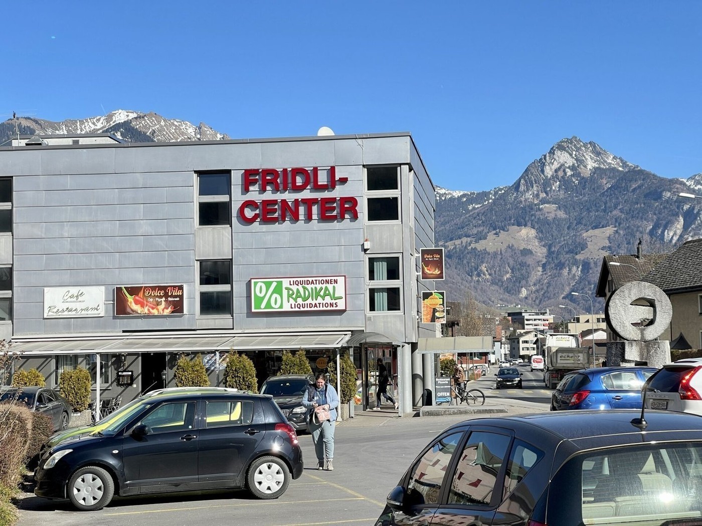 Commercial building with a sign reading 'FRIDLI-CENTER', multiple shops, cars parked in front, mountains in the background