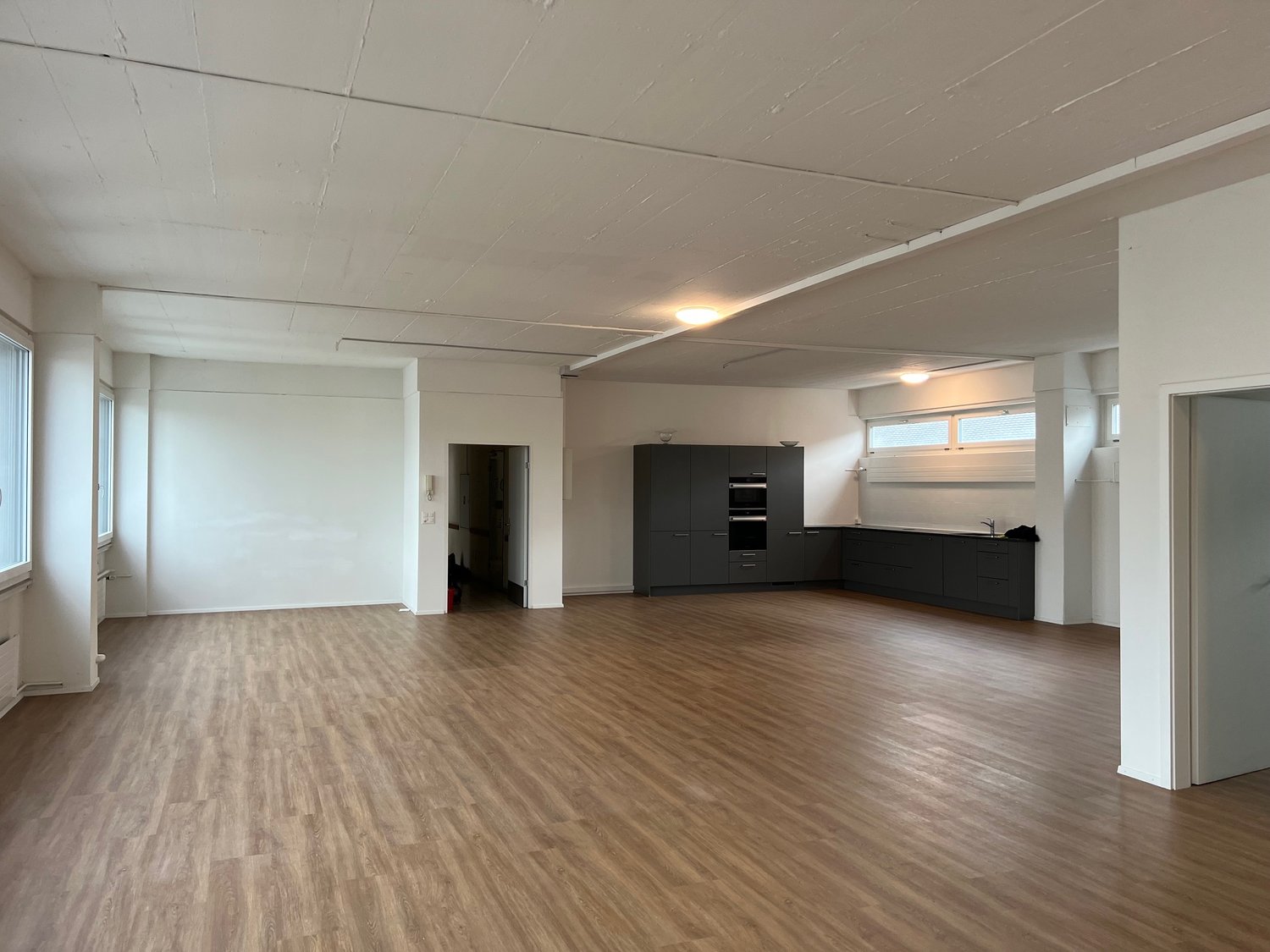 an empty, spacious room with wooden floors, white walls, and a kitchen area in the back