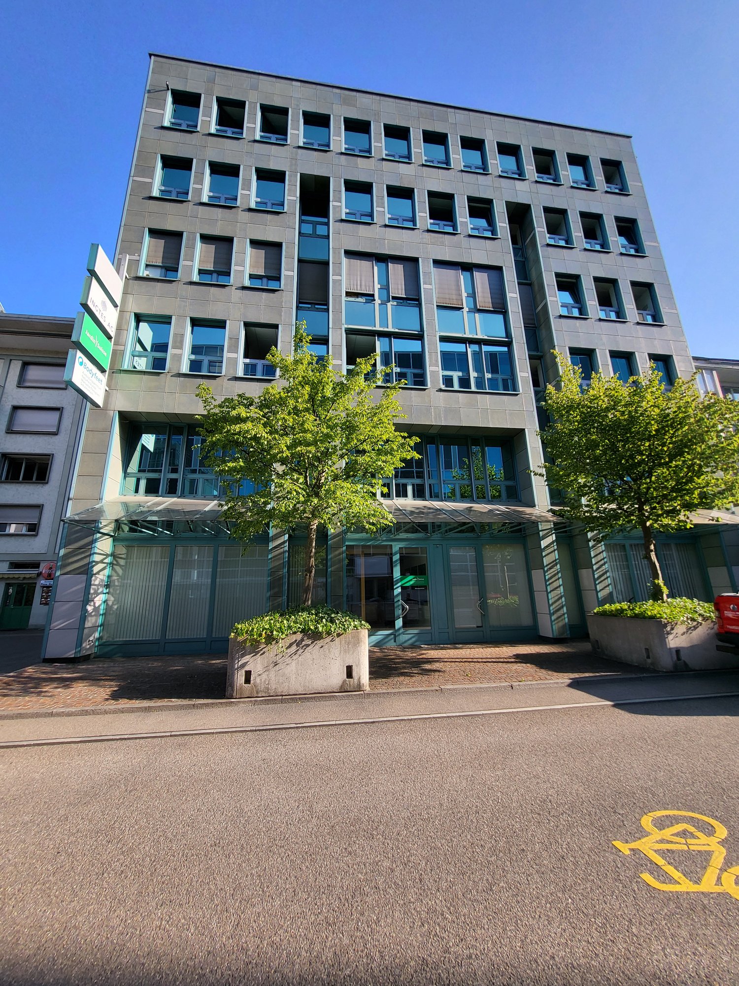 Multi-story office building with a modern, glass-and-concrete facade. The building has numerous windows, some of which appear to be balconies or terraces. There are trees and greenery surrounding the building, creating a pleasant, urban environment.