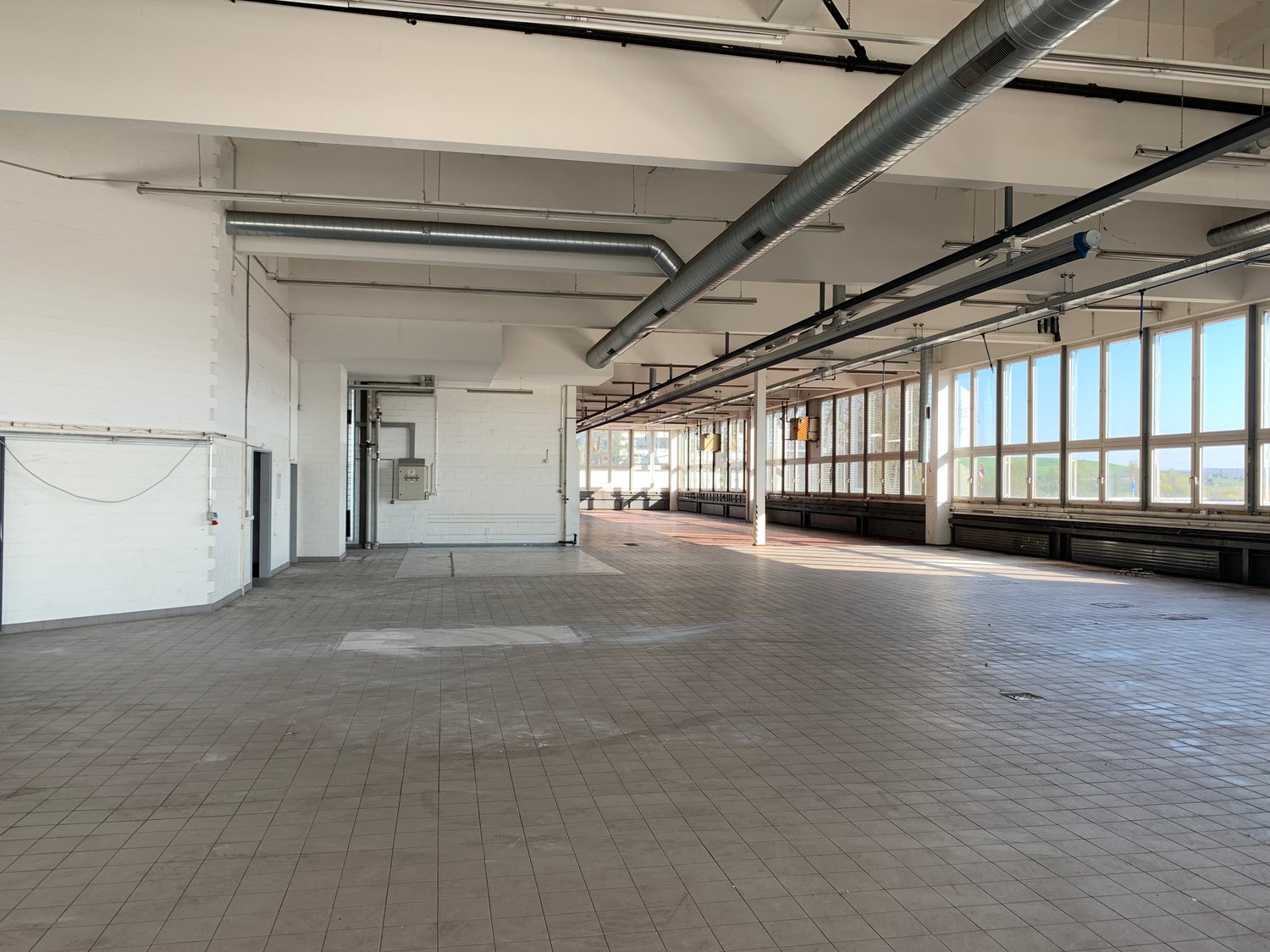 Large open commercial space with high ceilings, exposed ductwork, and tiled flooring. The space appears to be a warehouse or industrial facility with multiple windows along one side providing natural light. There are no visible furnishings or equipment, s