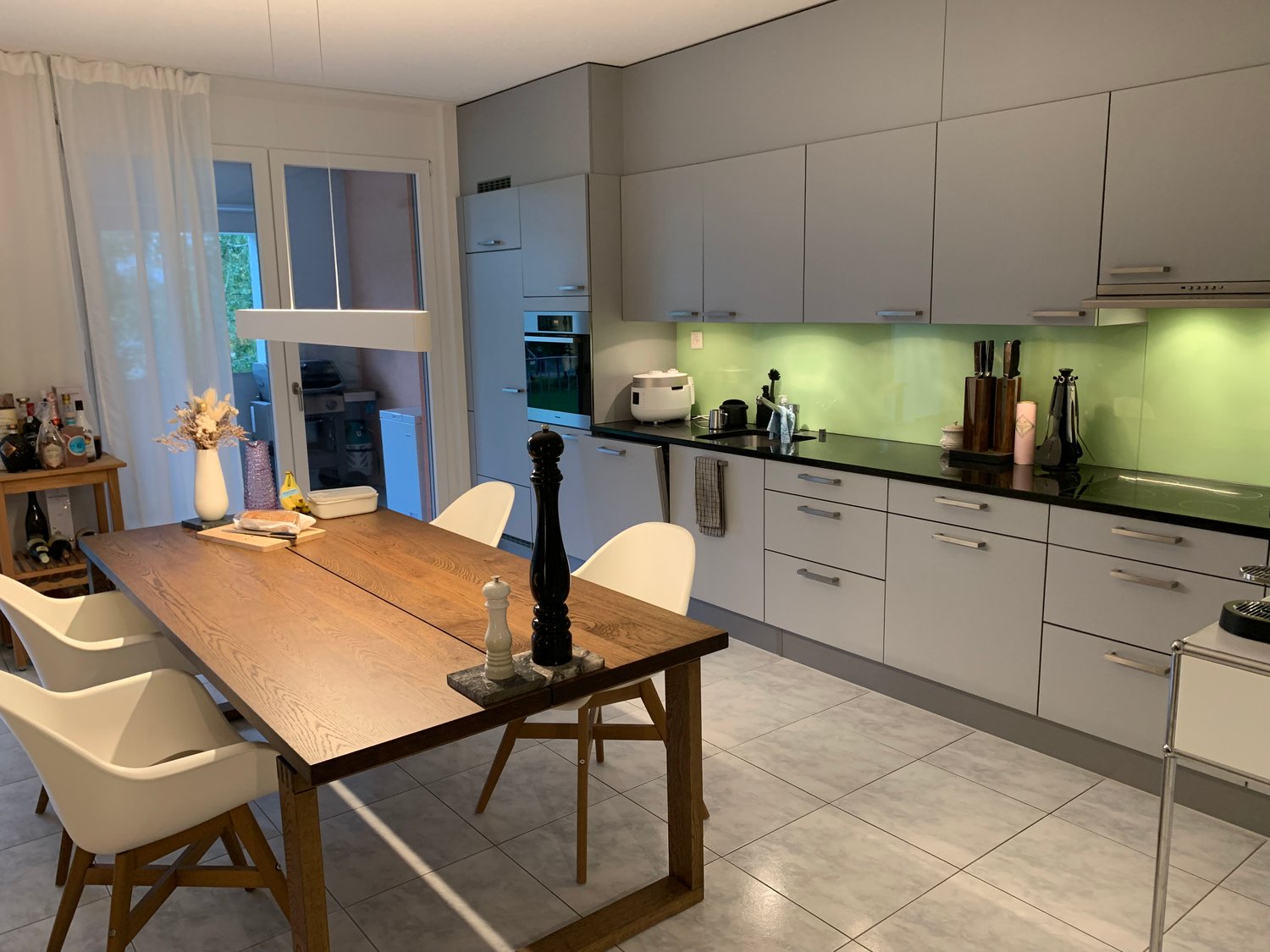 Kitchen and dining area, wooden dining table, white chairs, modern kitchen design, built-in appliances, white cabinets, black countertops, green backsplash, island with seating, breakfast bar.