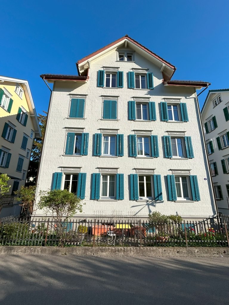 Apartment building, white exterior, multiple windows, fenced front yard, greenery