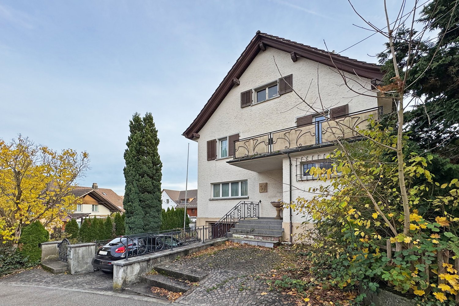 House with brown roof, white walls, brown shutters, balcony with black railings, steps to the entrance, car parked in front