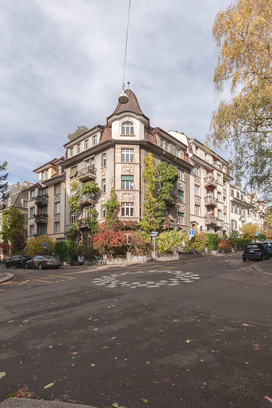 Multi-story building with balconies, garden, large front area, multiple parking spaces visible