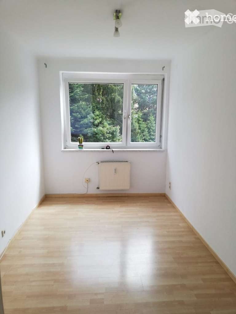 Spacious bedroom with large windows overlooking a lush, green outdoor area. The room has hardwood flooring and a radiator installed on the wall.