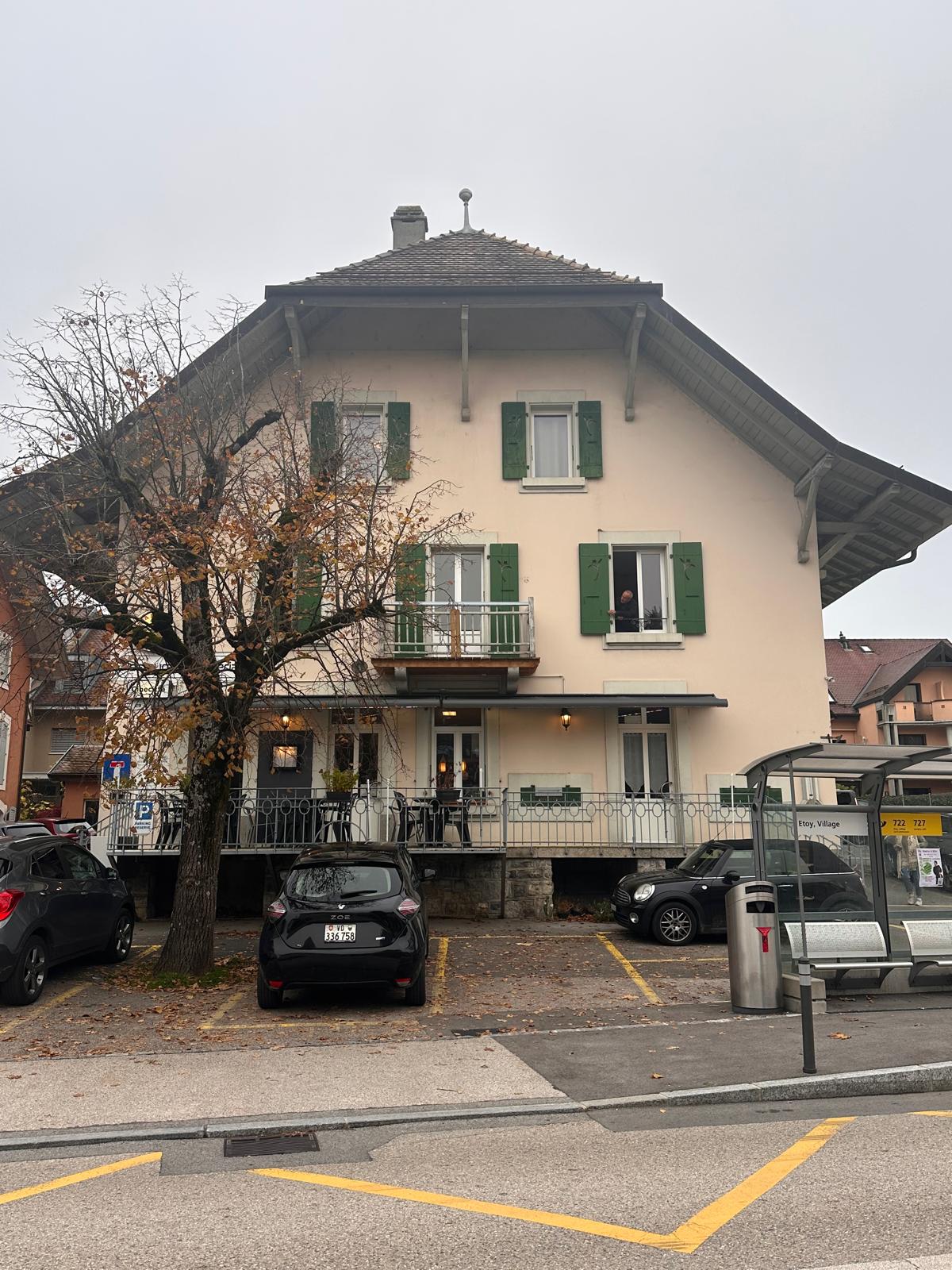 Two-story building, beige exterior, green shutters, wooden porch, multiple windows, front yard with trees, black car parked, nearby parking lot, trash can.