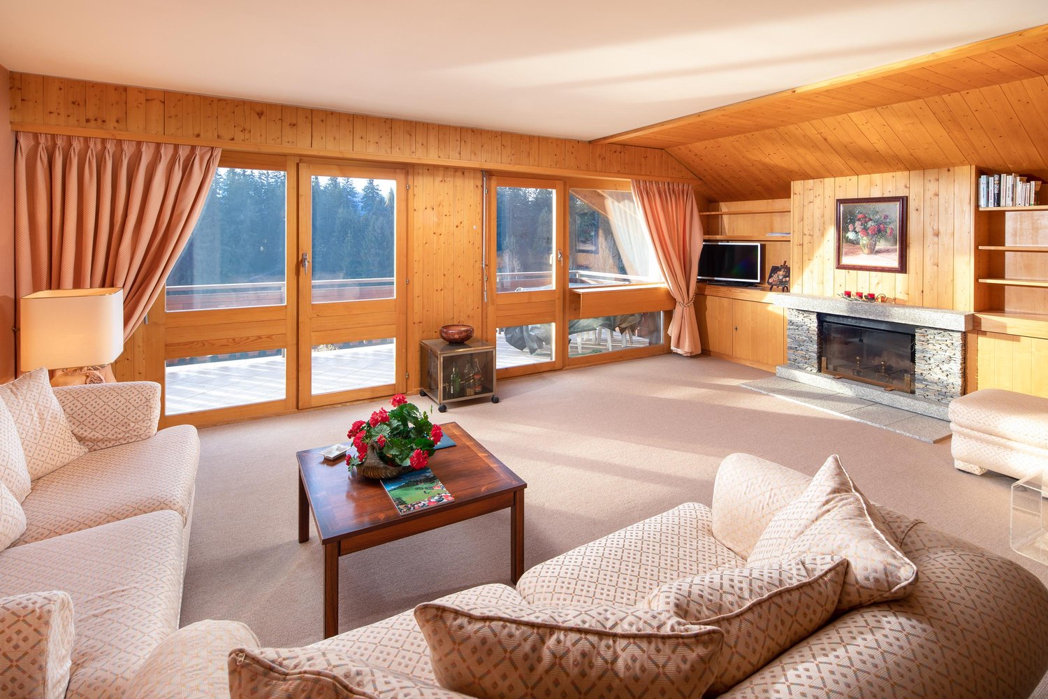 living room with fireplace, large windows, wooden walls, couches, coffee table, ottoman, lamp, shelves, TV
