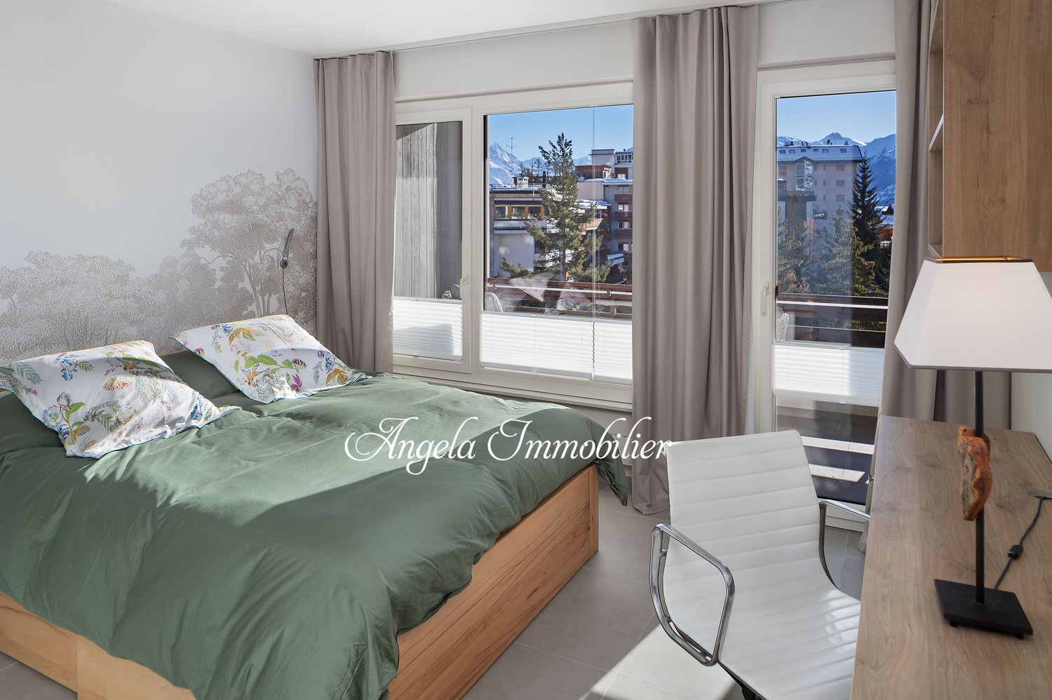 large bed, green linens, pillows, gray curtains, white walls, desk, chair, lamp, balcony, mountain view