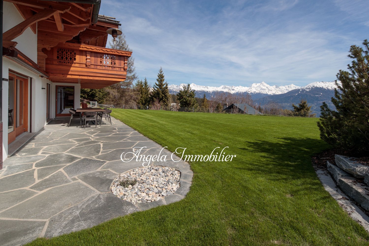 2-story wooden chalet-style house with a large balcony, surrounded by a well-maintained lawn and landscaping. The house has a stone patio in the foreground and offers a scenic view of snow-capped mountains in the background.