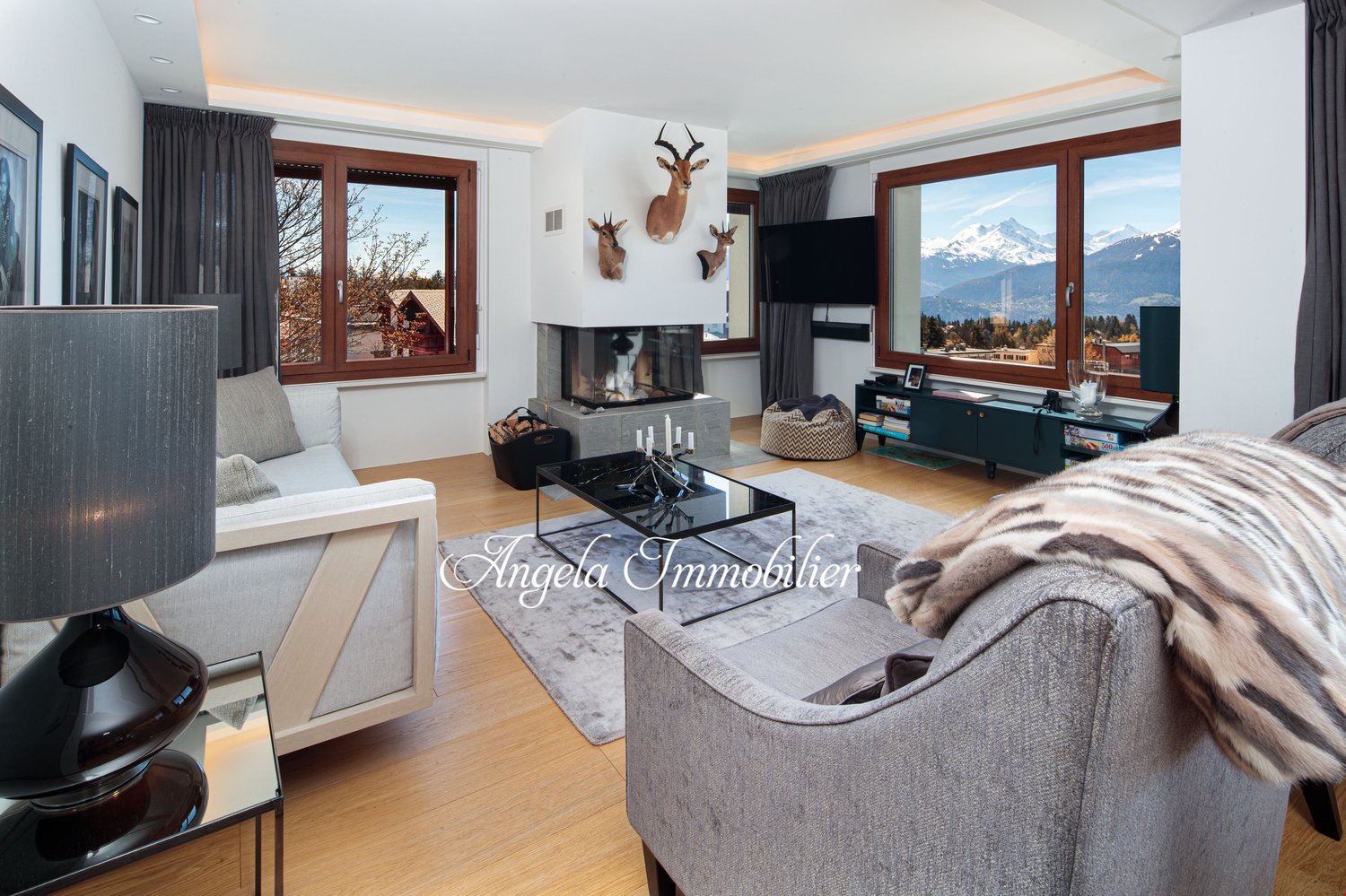 fireplace, 3 deer heads mounted on wall, 3 windows, 2 couches, gray area rug, glass table, gray armchair with fur blanket
