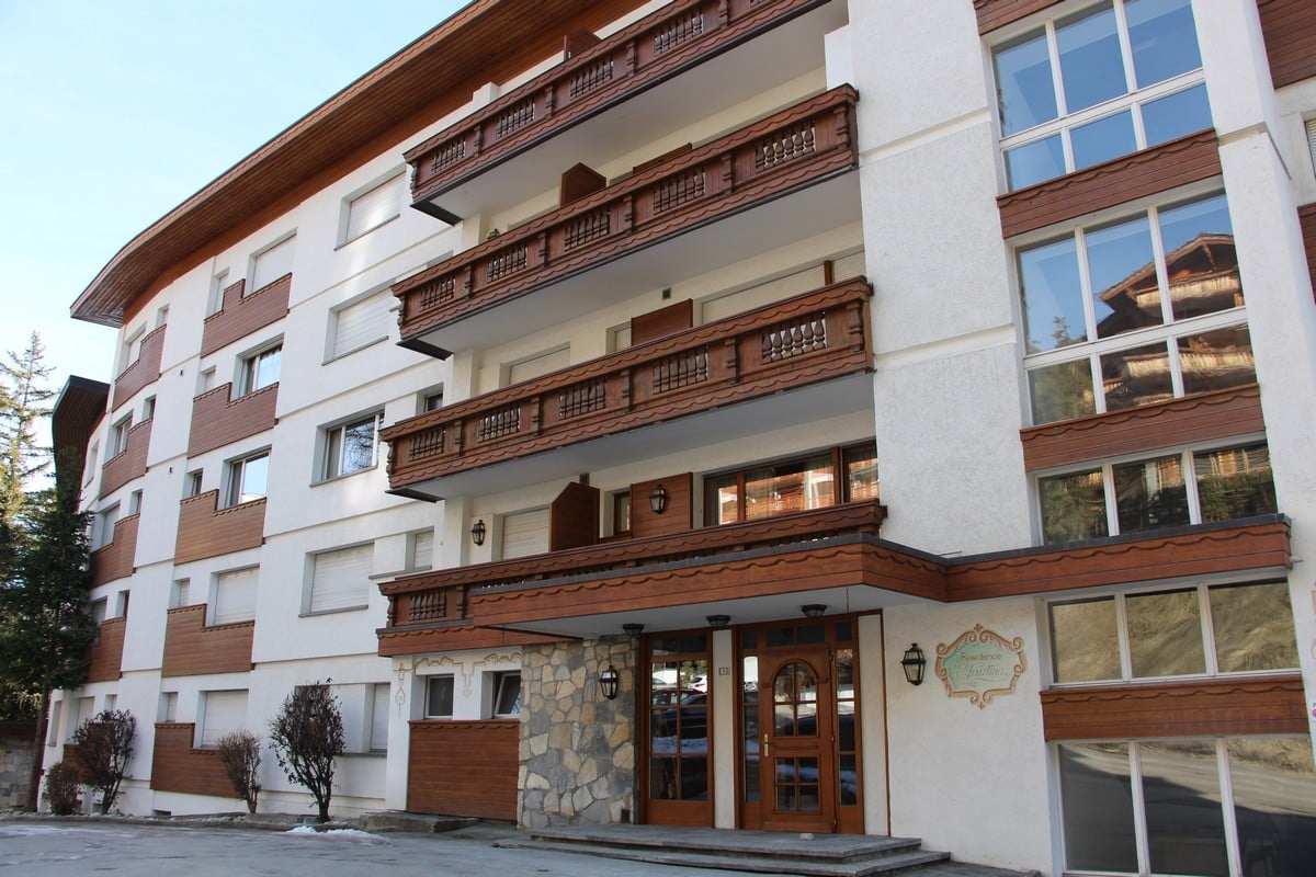 White and brown facade, 3 floors, large balconies with wooden railings, multiple windows, double doors at the entrance