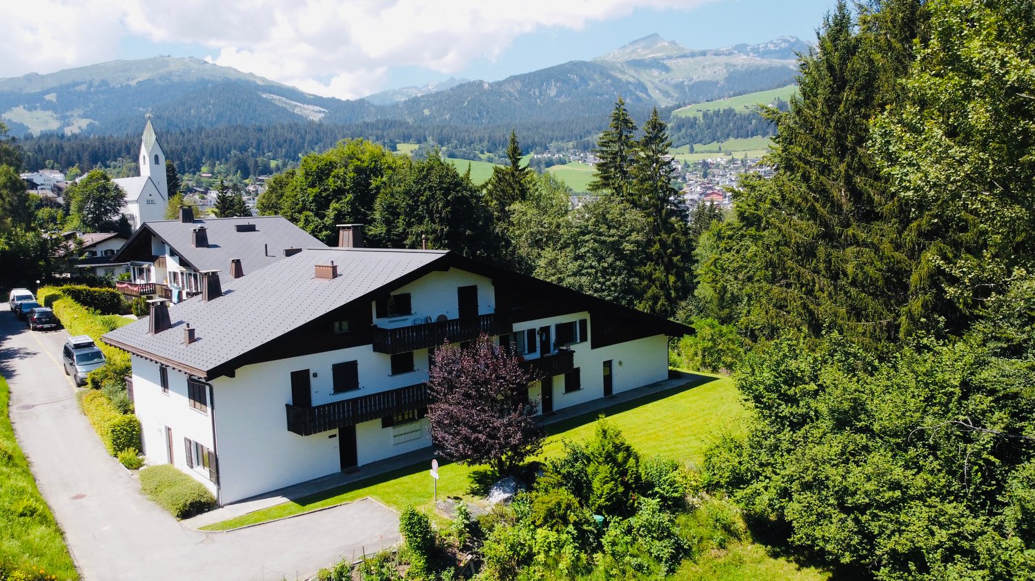 A multi-story residential building with a church steeple in the background, surrounded by lush green trees and mountains in the distance. The building has balconies and a tiled roof.