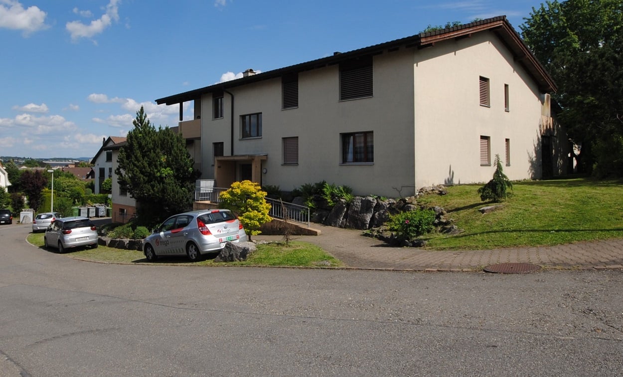 White, two-story house with a balcony and a driveway leading to the entrance, several cars parked nearby, shrubs, trees, and a grassy area in front.