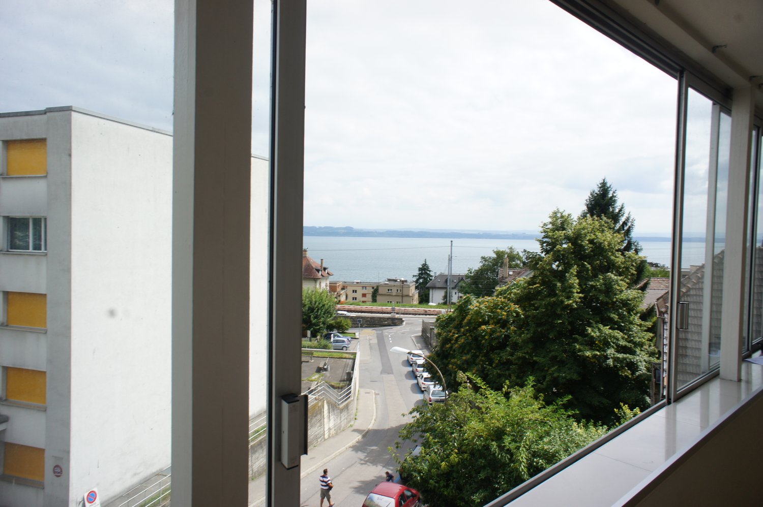 View from the window of a street with cars parked, a person walking, and a building with yellow shutters. In the distance, there is a body of water and a mountain range.