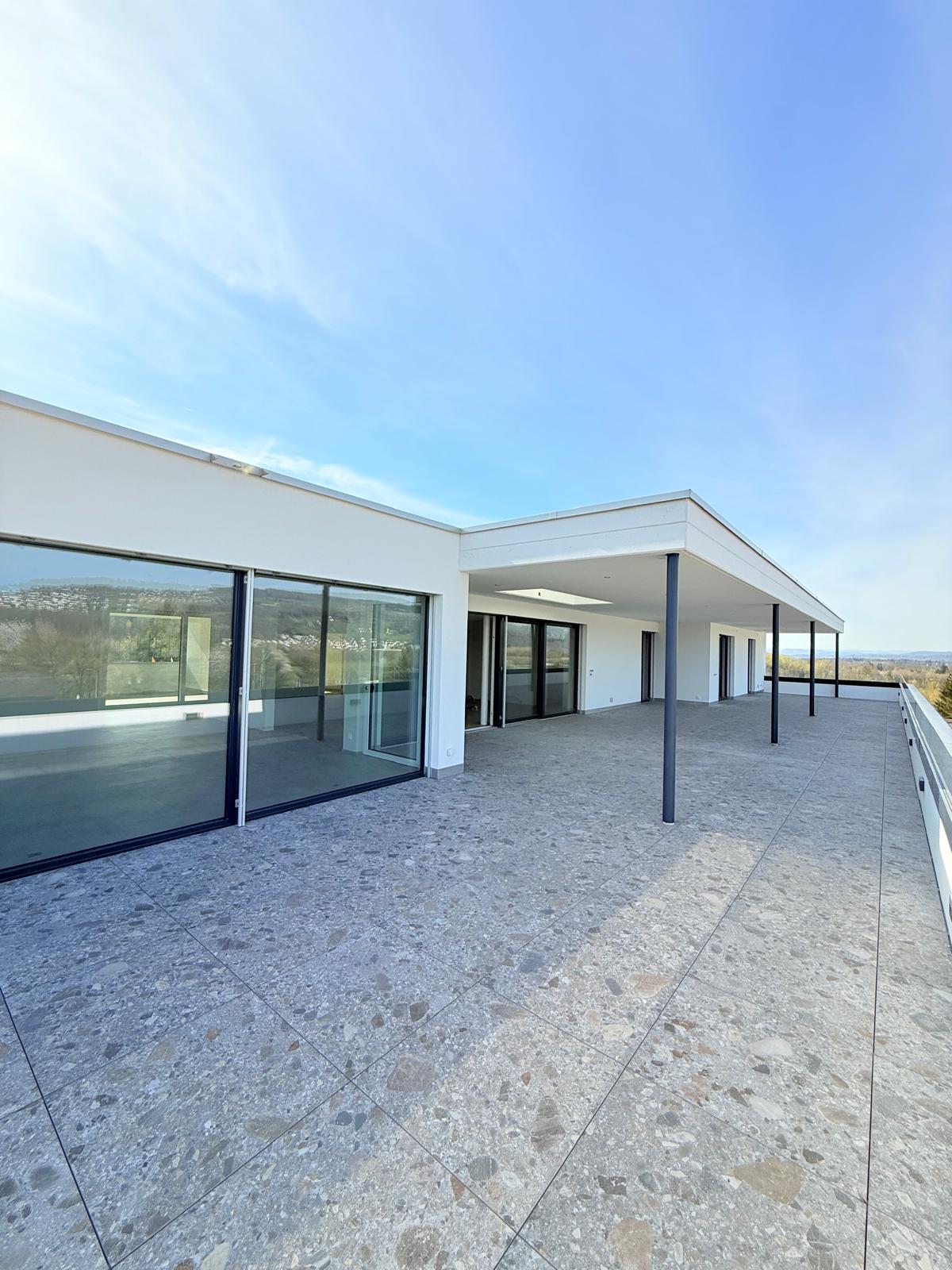 Modern two-story house with large glass windows, a covered patio or balcony area, and a tiled outdoor terrace.