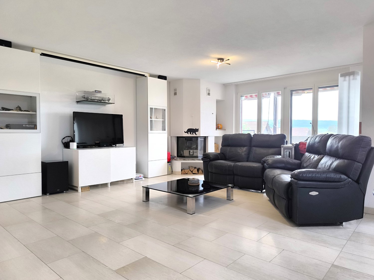 A living room with a modern design. It features a leather sofa, black glass coffee table, white cabinets, a mounted TV, and large windows with curtain. The floor is covered with white tiles. There is a fireplace at the back of the room with firewood stora