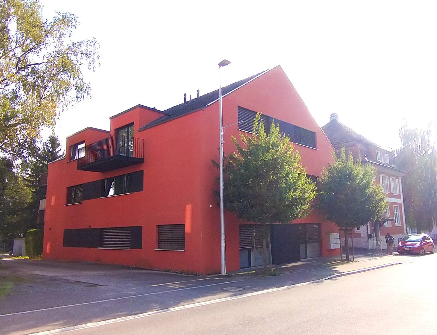 red building with black trim, multiple balconies, glass windows, trees, parked red car