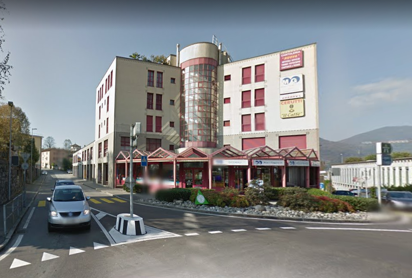 A multi-story building with a modern architectural design, featuring glass windows and red shutters. The building has a signboard with the text 'Il Caffe' and other advertisements. There are cars driving on the road in front of the building, and a pole wi