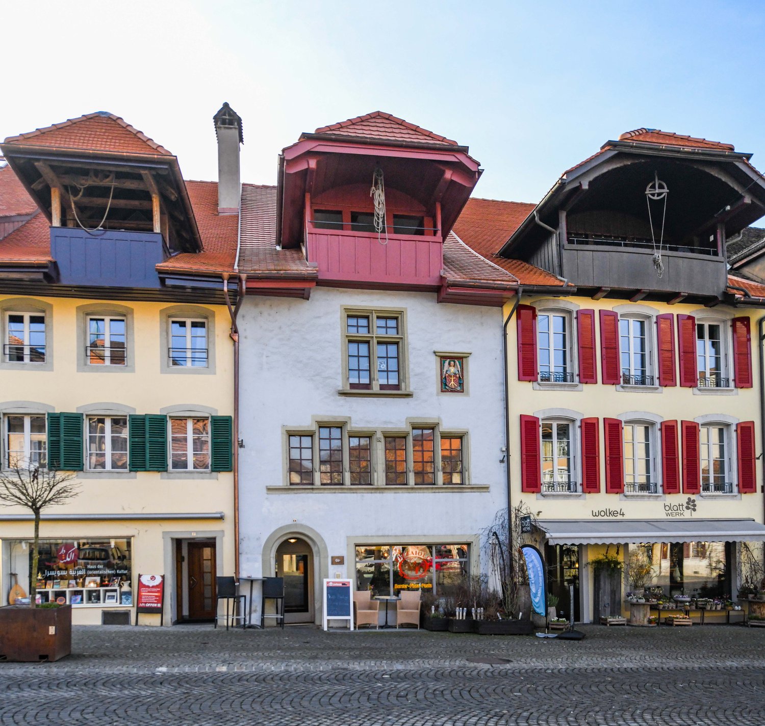 The image shows a row of traditional, colorful buildings with red tile roofs, wooden shutters, and decorative architectural elements. The buildings appear to be located in a town or city setting, with a cobblestone street in the foreground. The buildings