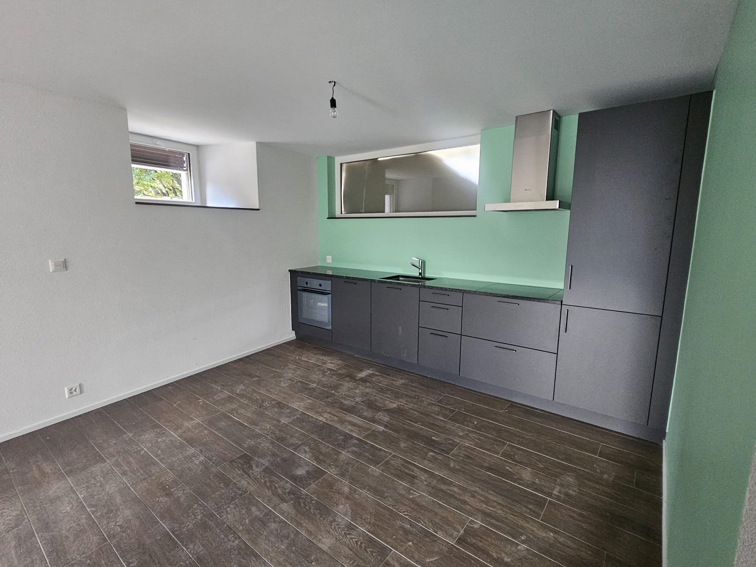 An empty kitchen with wooden floor and green wall, an oven below the sink, and cabinets on the wall.