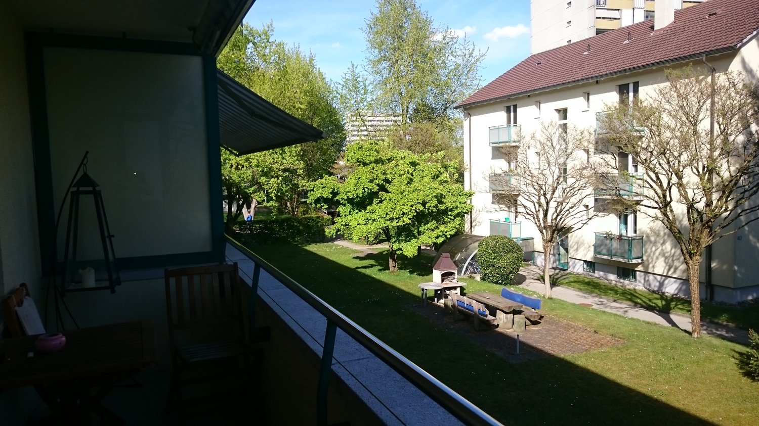 Balcony overlooking a green garden with trees, shrubs, and a small play area. The building has multiple floors and apartments with balconies.