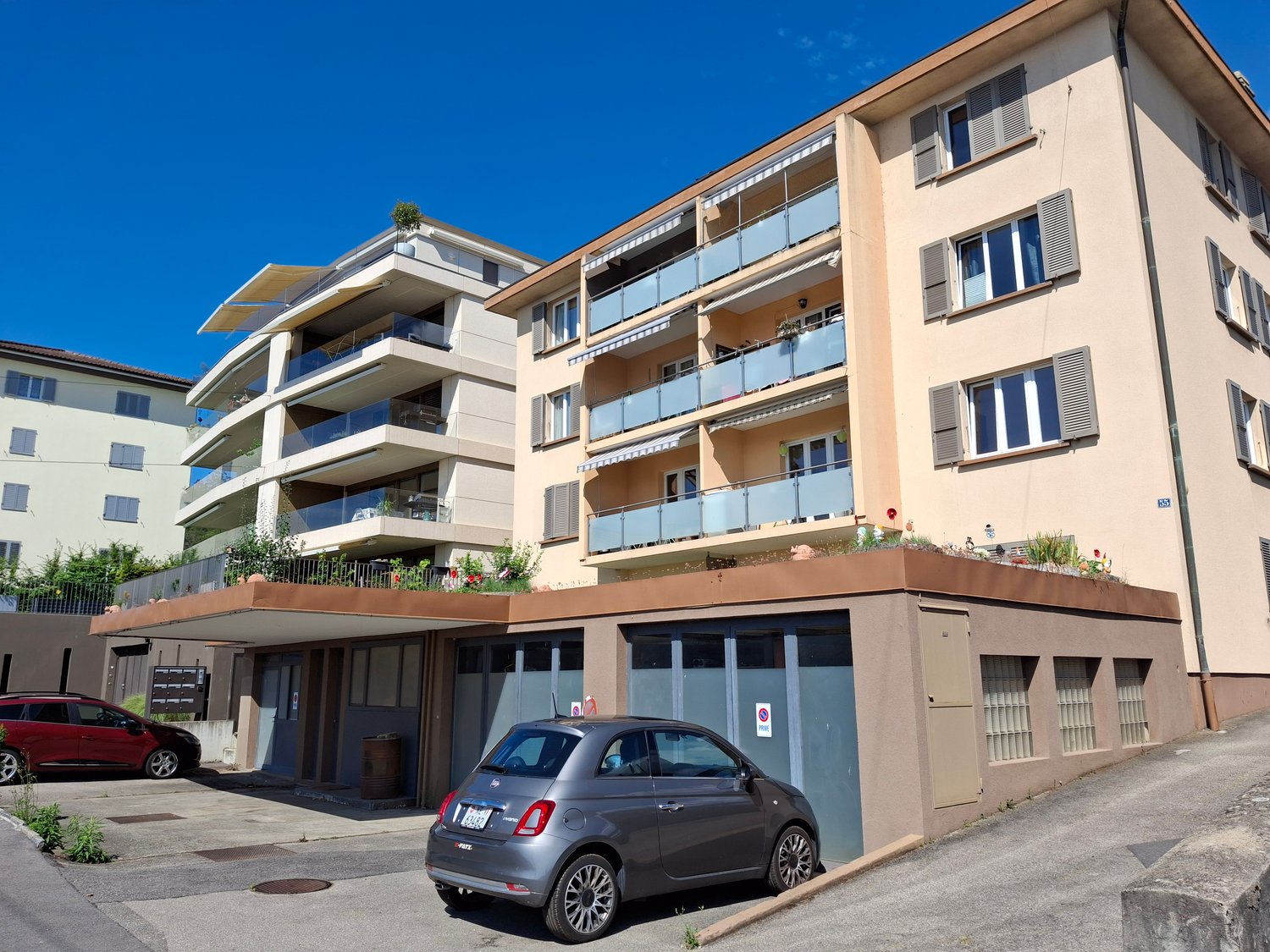 Three-story apartment building with balconies, parking, and a ground-floor garage. The ground floor has plants on the roof terrace and two cars parked.