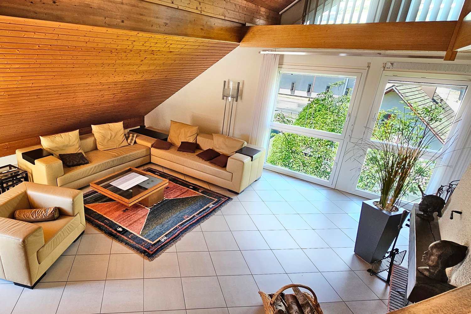 tiled floor, beige leather sofas, L-shaped sofa, wooden ceiling, coffee table, wooden rug, lamps, large windows, indoor plants, fireplace