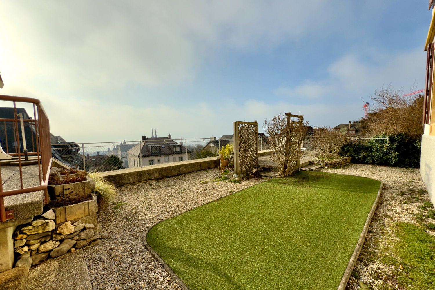 small garden with gravel and green lawn, stone wall, metal railing, view of the city