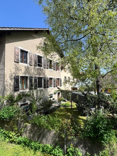 2 story building, beige colored, multiple windows, wooden shutters, garden with a tree, plants and shrubs, a small fenced area, and a wooden fence