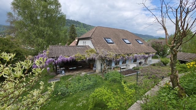 Two-story house with a tiled roof, surrounded by lush greenery and flowering plants. The house has several dormer windows on the upper floor and a raised ground floor. There appears to be a balcony or terrace on the upper level.