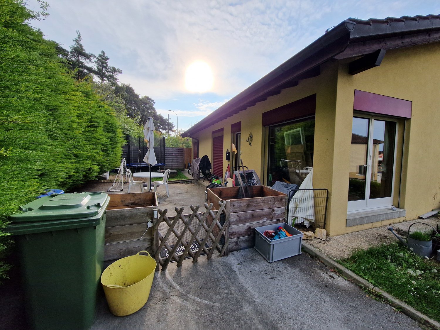 Single-story house with a yellow exterior, a slanted roof, and a covered patio or balcony area. There is a garage or storage area visible, and various outdoor items and equipment are present in the backyard.