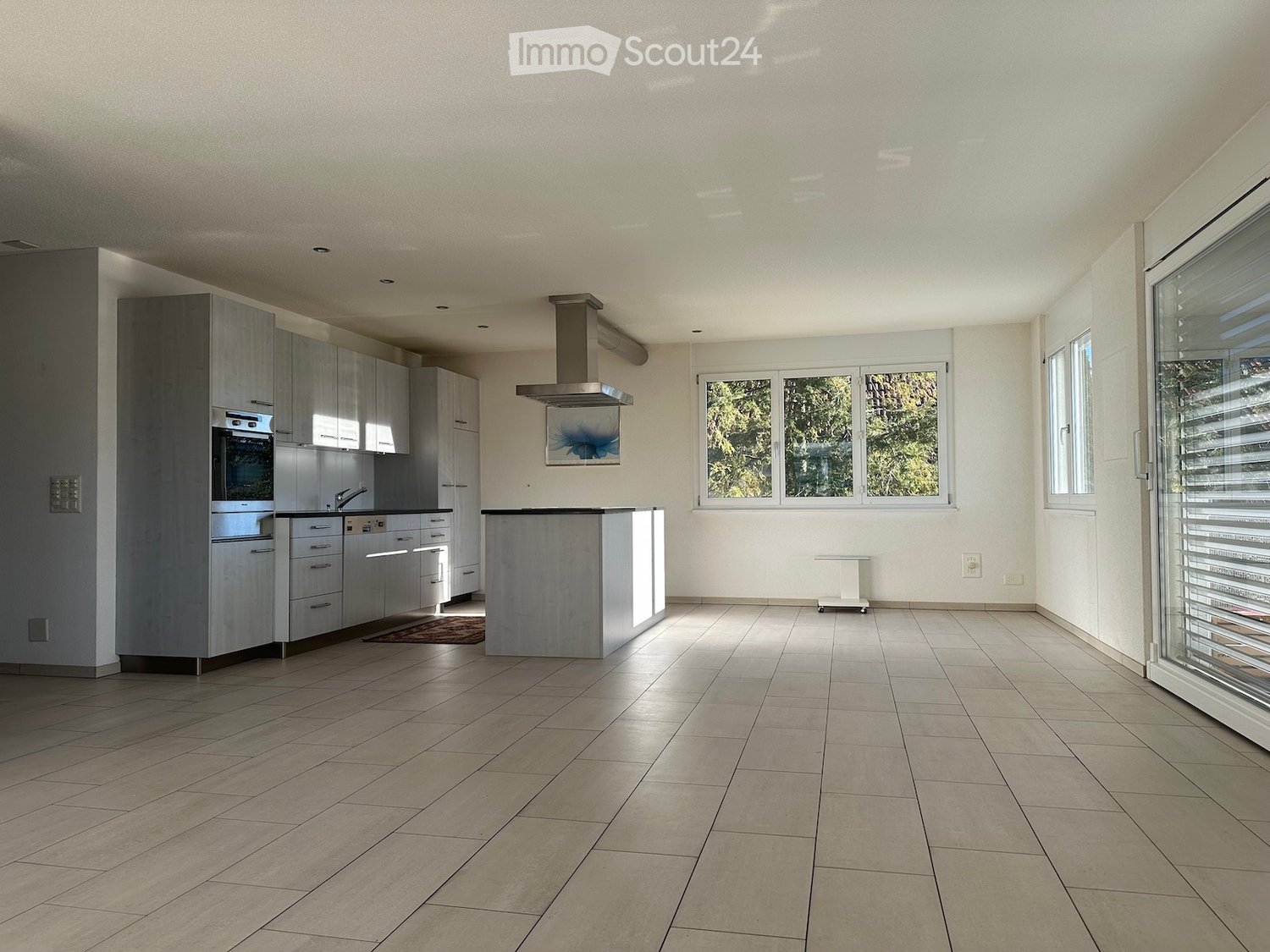 Spacious open-plan living area with modern kitchen, large windows, and tiled flooring throughout. The kitchen features grey cabinetry and stainless steel appliances, including a dishwasher.