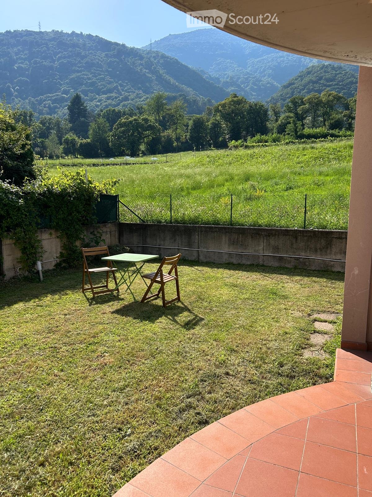 green table and chairs on a lawn, tiled floor, overlooking a grassy field and mountains in the distance