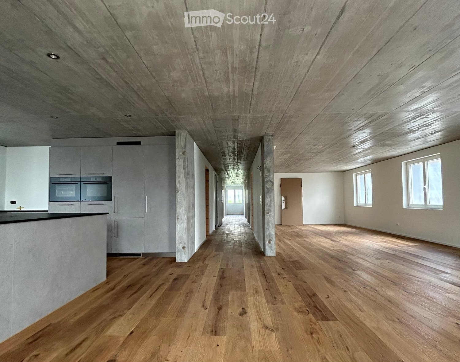 empty hallway with hardwood floor, white walls, exposed concrete ceiling, multiple windows, kitchen appliances