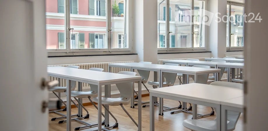 Empty classroom with tables and chairs arranged in rows, large windows facing the building.