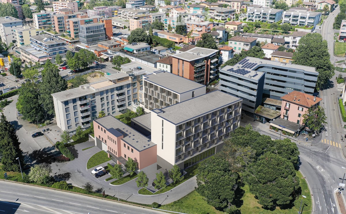 An apartment building with multiple floors and windows, solar panels on the roof, surrounded by trees and grass, a parking lot with a road in front.