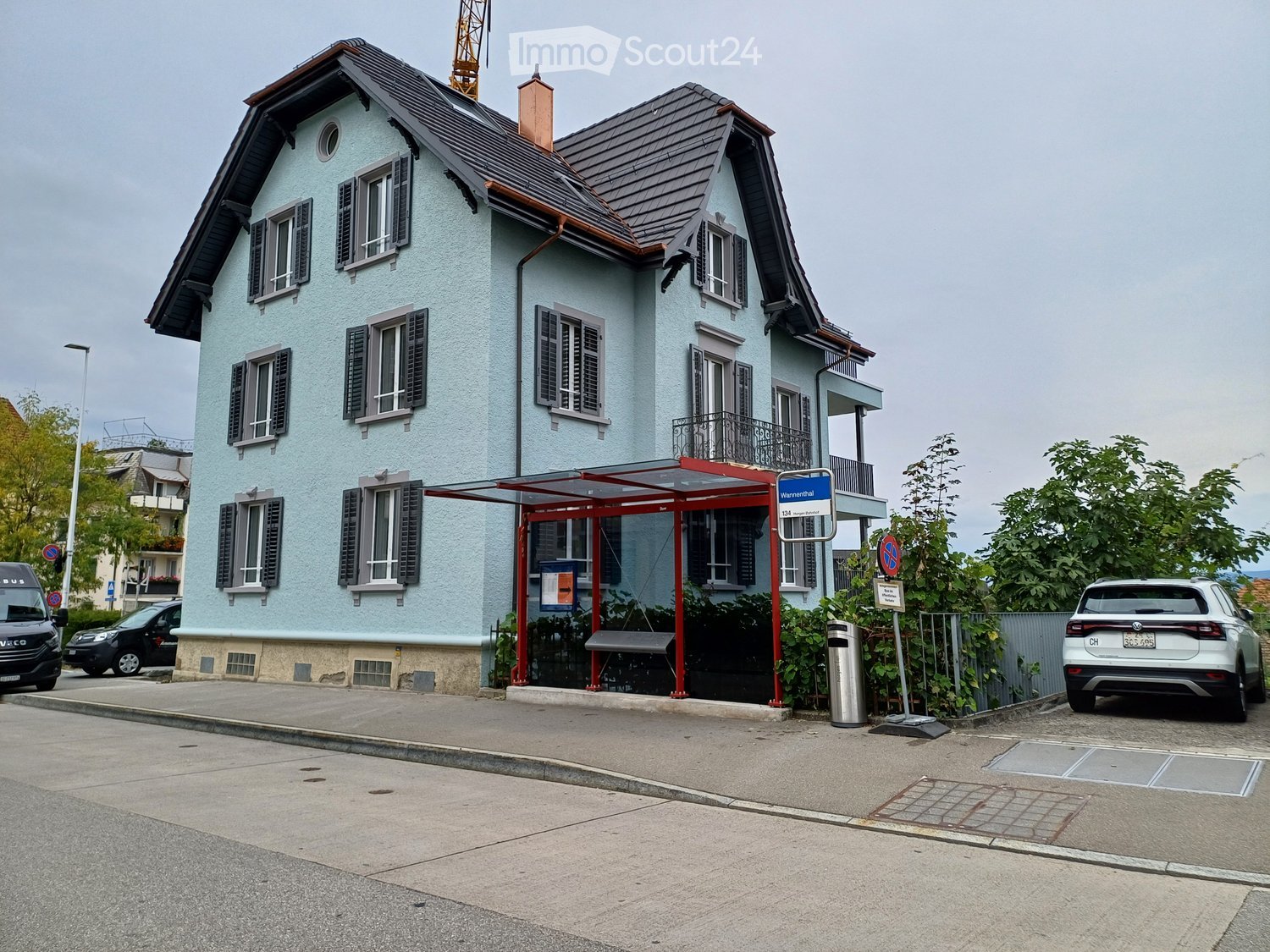 Large apartment building, 3 floors, light blue exterior, green roof, black exterior trim, windows with black shutters, entrance sheltered by a canopy, bus stop in front, sidewalk, street, parked cars, greenery