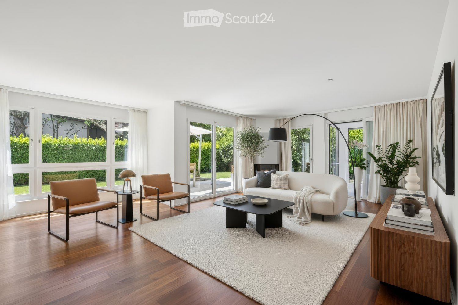 open plan living room, white walls, hardwood floors, large windows with glass doors leading to the garden, white couch, round black coffee table, beige rug, arc floor lamp, side table with a lamp, plant, wood side table with books, large white painting on