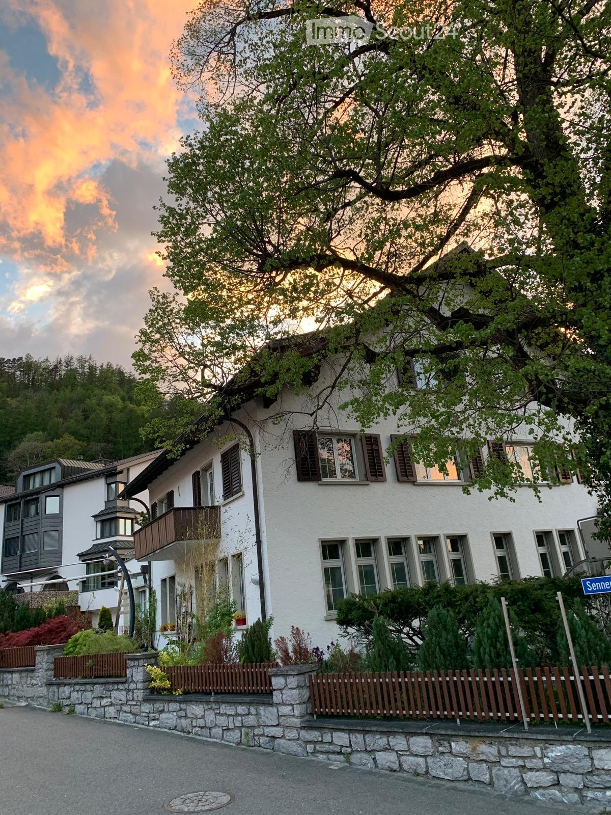 A traditional white house with brown shutters, a balcony, well-maintained garden with trees, shrubs and fences