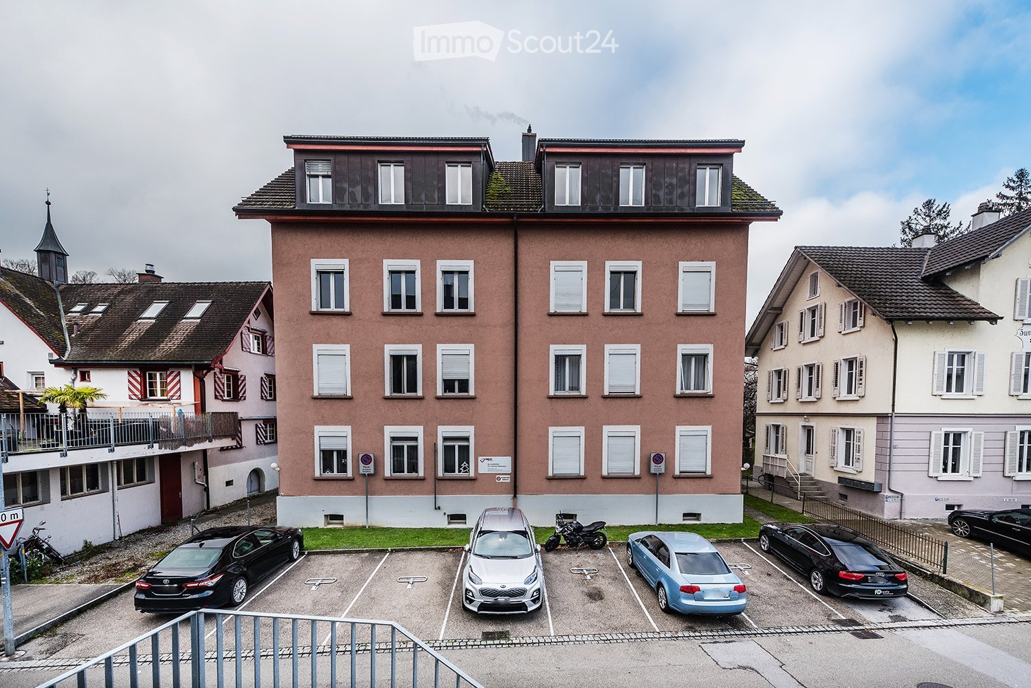 Apartment building with multiple windows, ground floor parking spaces, and additional structures in the background.