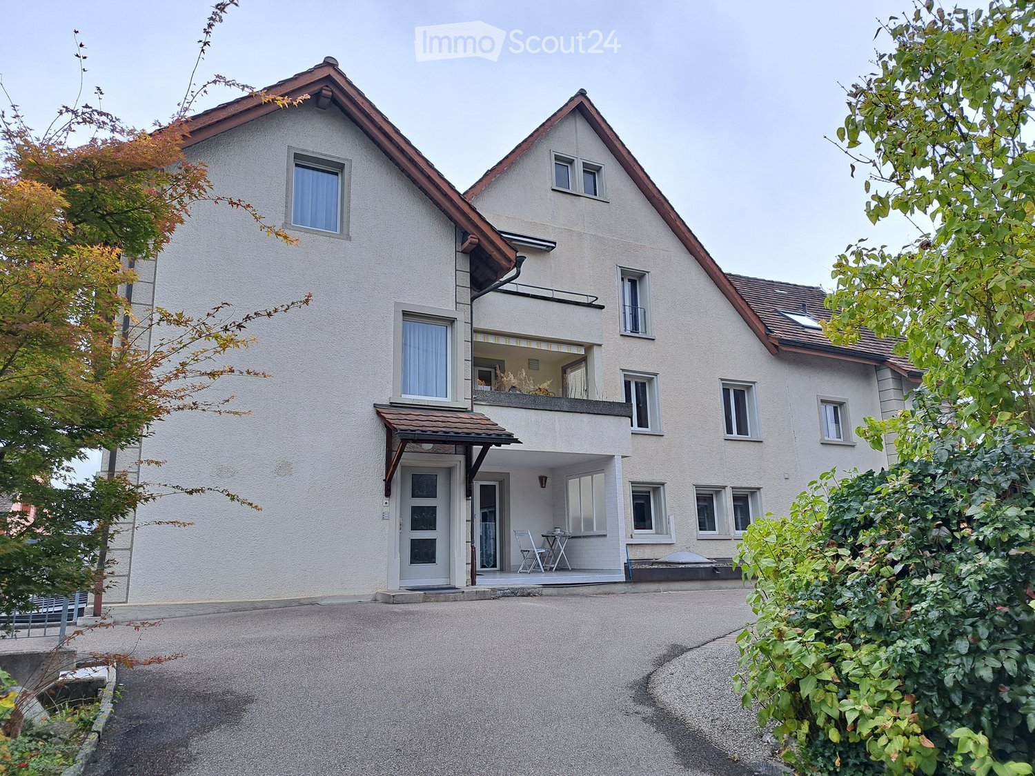 A multi-story building with a white exterior and brown roof, featuring multiple windows, a balcony on the second floor, and a small patio with a table and chairs in front of the entrance.