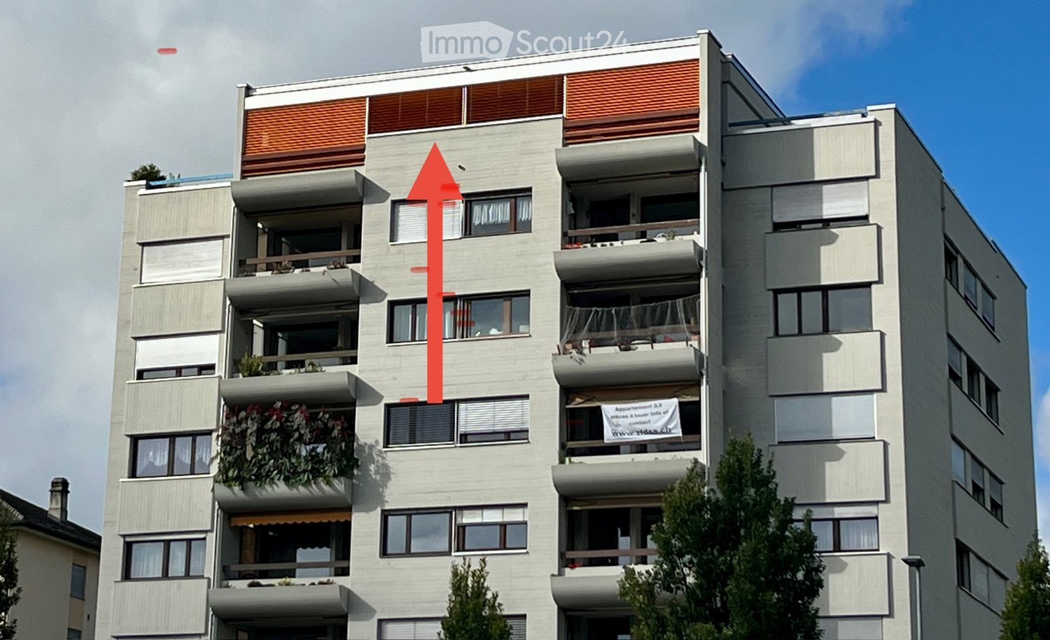 Apartment building, multiple floors, grey exterior, windows with shutters, some balconies with plants, white sign with red arrow pointing upwards