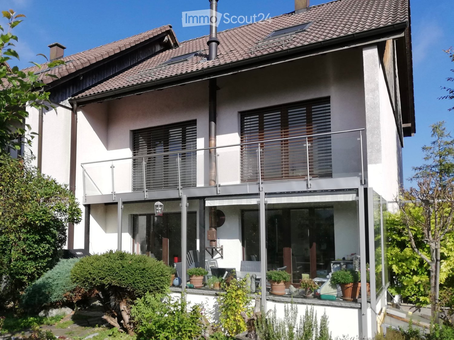 Modern two-story house, white exterior, tiled roof, large balcony, large windows, outdoor seating area, fenced garden, several plants